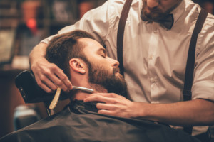 Making hair look magical. Close-up side view of young bearded man getting shaved with straight edge razor by hairdresser at barbershop